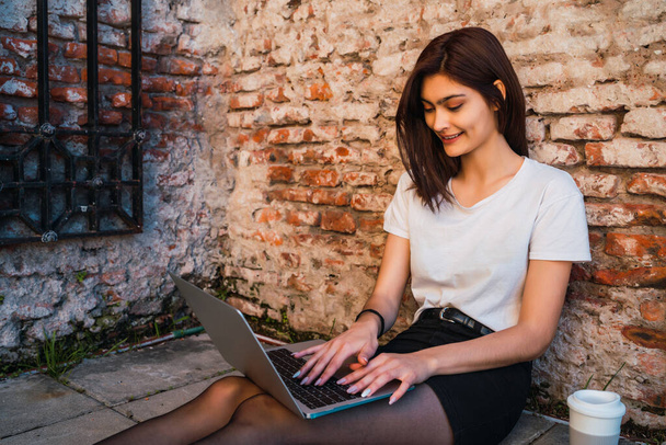 Portrait of young latin woman relax and using laptop while sitting outdoors against brick wall. Urban concept. - Fotografie, Obrázek