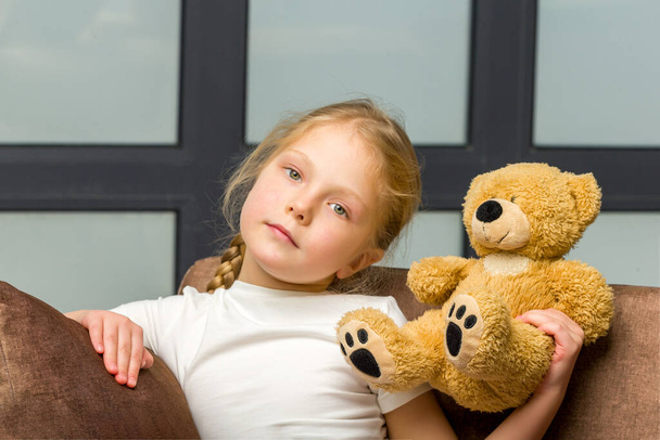 Little girl on sofa hugging teddy bear. - 写真・画像