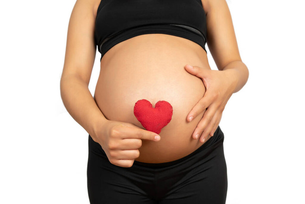 Close-up of pregnant woman holding heart sign on belly against white background. Pregnancy, motherhood concept. - Фото, зображення