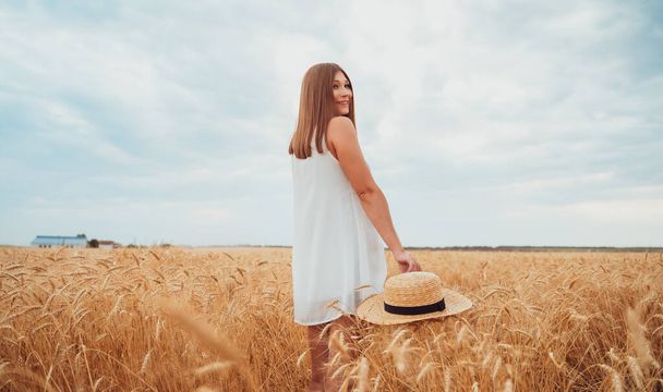 A young beautiful model in a white dress, inspired by nature, walks through a wheat field and smiles happily, holding a straw hat in her hands, which lies on the ears of ripe rye. - Foto, imagen