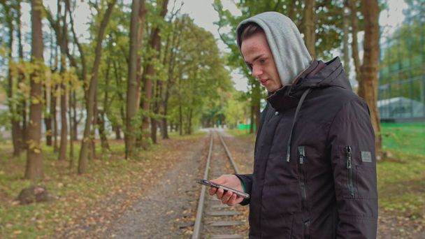 Gezicht op een millennial hipster, het openen en scrollen van het telefoonscherm. Jongeman met behulp van een smartphone tijdens het lopen op straat in het park. Een jongeman met een kap kijkt naar de telefoon. - Foto, afbeelding