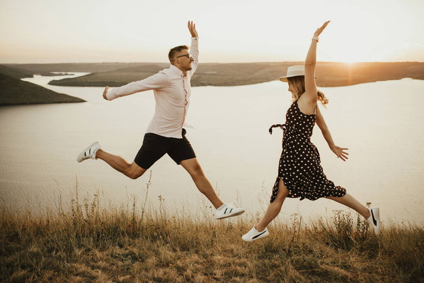 man and woman in love jumping on mountain above large lake by the sea at sunset - Foto, imagen