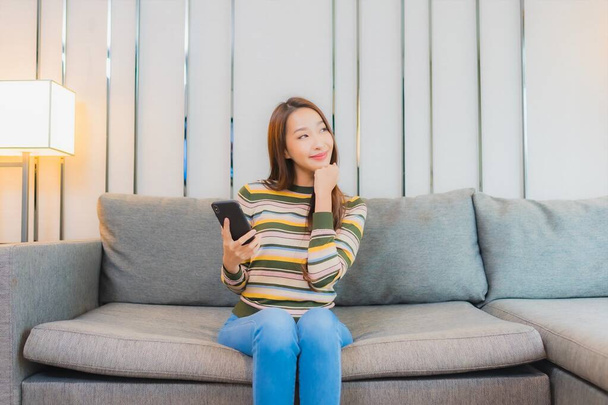 Πορτρέτο όμορφη νεαρή Ασιάτισσα γυναίκα χρησιμοποιούν έξυπνο κινητό τηλέφωνο στον καναπέ στο εσωτερικό του σαλονιού - Φωτογραφία, εικόνα