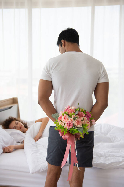 Family relationship and happy anniversary concept. Young romantic man holding the bouquet of flower hiding his back for surprising his wife laying on bed - Photo, Image