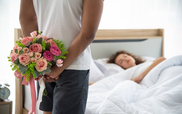 Family relationship and happy anniversary concept. Young romantic man holding the bouquet of flower hiding his back for surprising his wife laying on bed - Фото, зображення