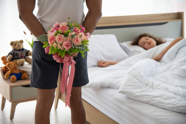 Relacje rodzinne i koncepcja szczęśliwej rocznicy. Młody romantyk trzyma bukiet kwiatów ukrywając plecy za zaskoczenie żony leżącej na łóżku - Zdjęcie, obraz