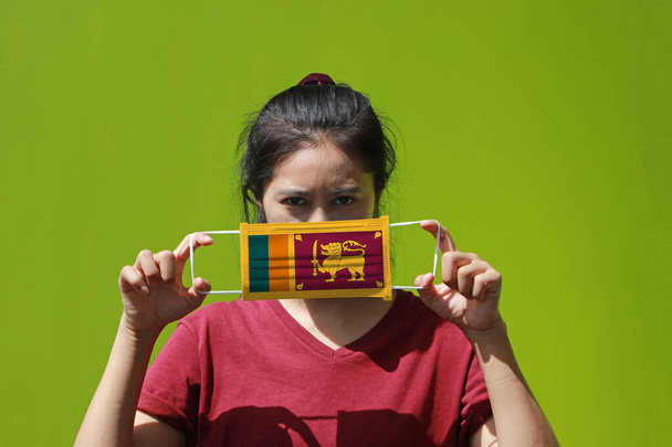 Una mujer y máscara higiénica con patrón de bandera de Sri Lanka en la mano y lo levanta para cubrir su cara sobre fondo de pared verde. Una máscara es una muy buena protección contra la diminuta partícula o la corona del virus. - Foto, imagen