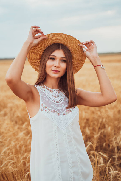 A young model in a white dress, inspired by nature, gently holds her straw hat on her head with beautiful hands, blurred background, portrait of a girl - Fotó, kép