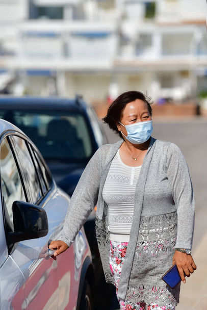 θέα ενός ήσυχο αναζητούν ώριμη ασιατική γυναίκα χρησιμοποιώντας μια μάσκα προσώπου στέκεται δίπλα σε ένα αυτοκίνητο και αρπάζοντας το χερούλι της πόρτας του αυτοκινήτου. Νέα έννοια της μετακίνησης και της ασφάλειας. - Φωτογραφία, εικόνα