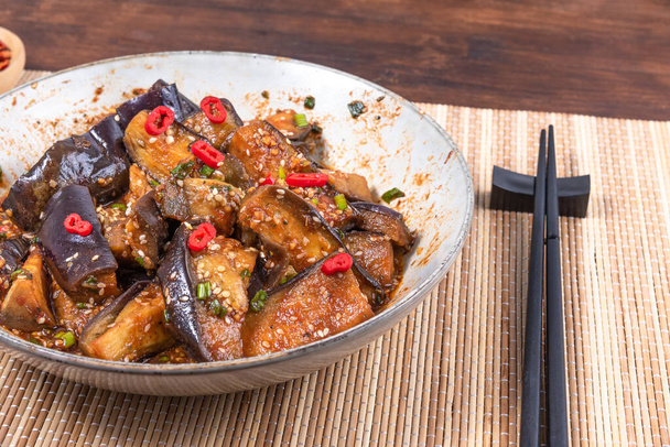 Korean spicy eggplant with spicy sauce and sesame seeds in a large bowl and chopsticks on a bamboo napkin on a rustic wooden table - Foto, immagini