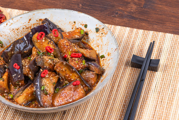 Korean spicy eggplant with spicy sauce and sesame seeds in a large bowl and chopsticks on a bamboo napkin on a rustic wooden table - Foto, afbeelding