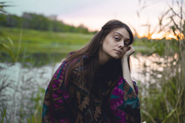 A young girl sits on the background of a sunset on the bank of the river in the reeds, leaning her hand to her face, an Indian poncho on a model, enjoying nature - Fotografie, Obrázek