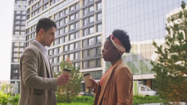  Seitenansicht eines gemischten Geschäftsmannes und einer schwarzen Geschäftsfrau, die vor einem modernen Business Center stehen und Kaffee aus To-Go-Tassen trinken, während sie über die Arbeit diskutieren - Filmmaterial, Video