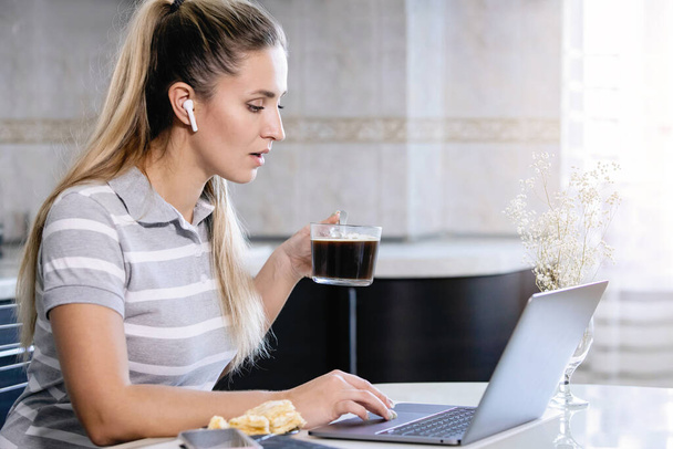 Kadın kahve içiyor ve masada dizüstü bilgisayarla çalışıyor. Tabakta pasta, günlük, bardak ve akıllı telefon var. Seçilmiş Odak. Coronavirus karantina ödevi, sosyal mesafe, özel girişimci. - Fotoğraf, Görsel