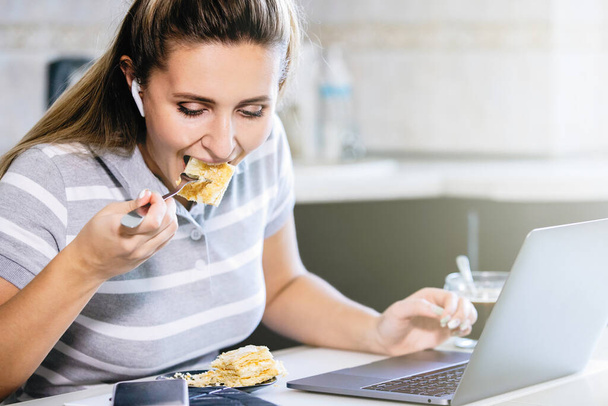 Woman at the table with laptop and cake at homework. Kitchen interior, diary, smartphone, wireless headphones. Epidemic, coronavirus pandemic. - Foto, Imagem