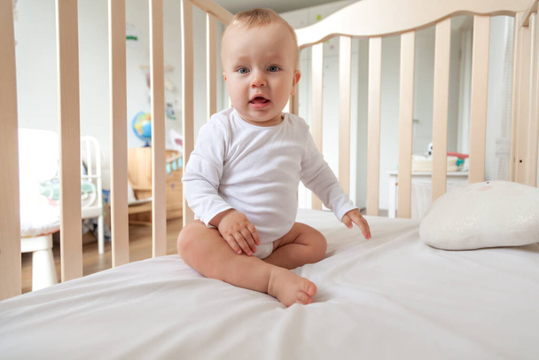 Szőke aranyos kis kék szemű baba fehér testöltözékben ül fából készült ágyban. - Fotó, kép