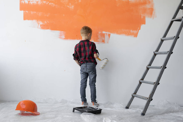 chłopiec stoi przy ścianie i maluje ją na pomarańczowo. Pomoc rodzicom. - Zdjęcie, obraz