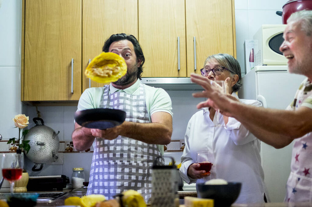Funny family in the kitchen cooking eggs and jumping them, surprised and fun expressions from senior mother and father while adult son jump the food - Photo, Image