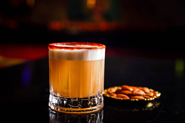 An orange cocktail in a rocks glass with a big ice cube, paprica crust rim, served with almonds - Fotó, kép