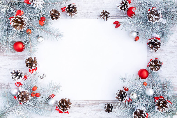 Elegante marco navideño con ramas de abeto cubiertas de nieve, conos y decoraciones navideñas en colores blanco y rojo. Hermosa plantilla de diseño de año nuevo con hoja blanca de papel en blanco para espacio de copia. - Foto, imagen