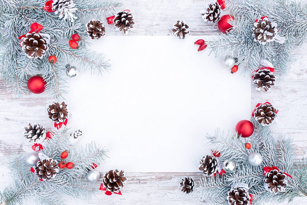 Fehér és piros karácsonyi keret vagy koszorú üres papír, fenyőtoboz és karácsonyfa dekorációk lapos feküdt a tetejére. Stílusos karácsonyi háttér hófödte lucfenyő ágak és másolás tér. - Fotó, kép