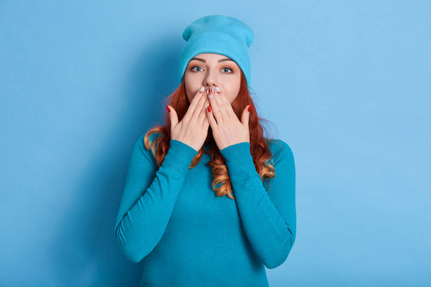 Stunned red haired woman with wavy hair, covers both hands on mouth, afraids of something,looks astonished, dressed in casual shirt, isolated over blue background, stands speechless indoor. - Фото, изображение