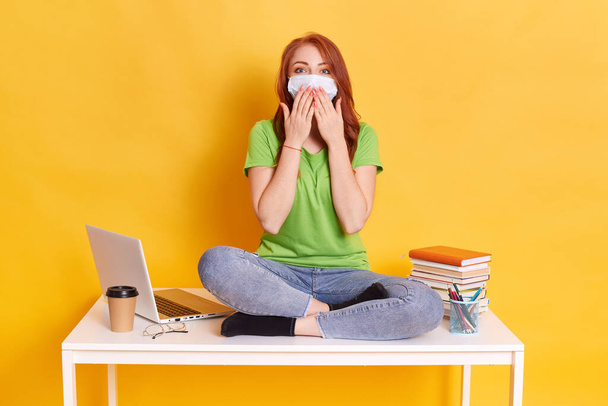 Photo of smart intelligent girl sits on table, preparing for exam or test, wears medical mask, green casual t shirt and jeans, works with lap top, isolated on yellow background, cover mouth with palms - Fotoğraf, Görsel