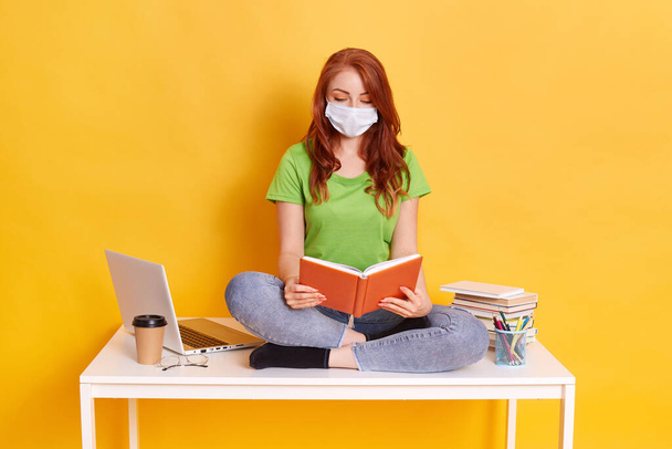 Photo of intelligent girl sitting table and studying, reading textbook, wearing green t shirt, jeans and medical mask over yellow color background, surrounded with books, lap top, take away coffee. - Fotó, kép