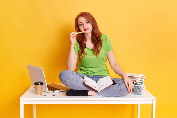 Young female student thinking about homework, red haired girl with yellow pan near her chin, wearing green casual t shirt and jeans, looks thoughtful while sitting on white table with crossed legs. - Foto, Bild