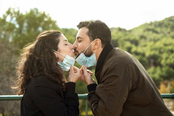Couple kisses outdoors with a surgical mask. Outdoor portrait symbolizing pandemic love - Foto, Imagem