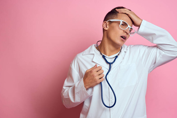 male doctor in a medical gown with a stethoscope around his neck on a pink background and glasses on his face - 写真・画像