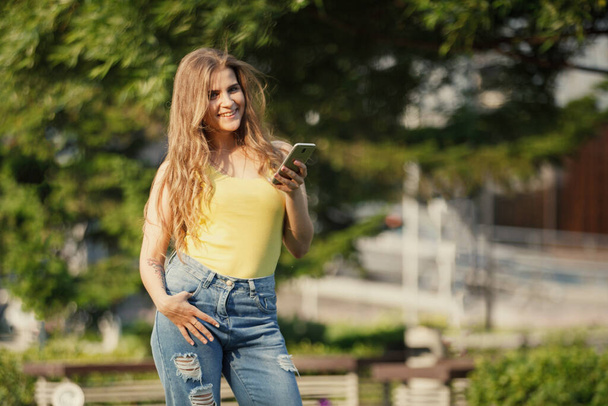 Donna con smartphone in t-shirt gialla e jeans in un parco cittadino. - Foto, immagini