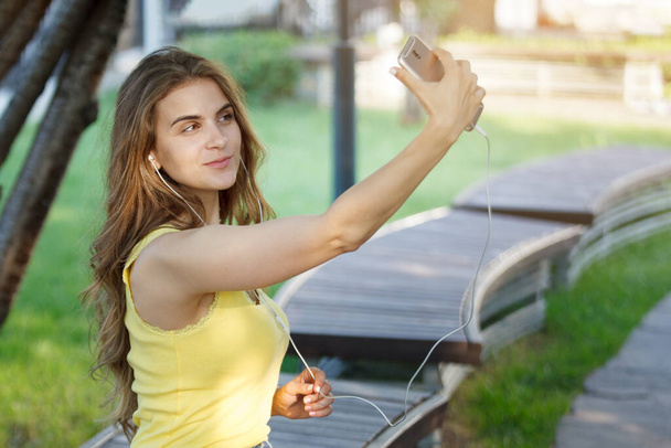 Mujer hace selfie con un teléfono inteligente en una camiseta amarilla y jeans en un banco en un parque de la ciudad. - Foto, Imagen