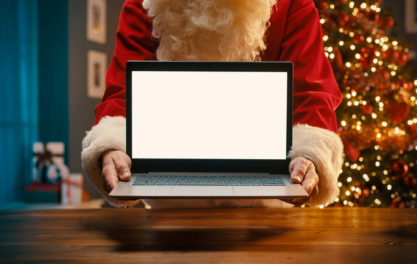 Santa Claus holding a laptop with blank screen, tree with lights in the background, Christmas and holidays concept - Photo, image