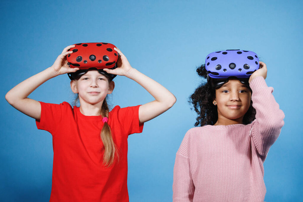 African American and blonde teenage girls play with virtual reality glasses. Computer games, blue background, emotions. - Photo, Image