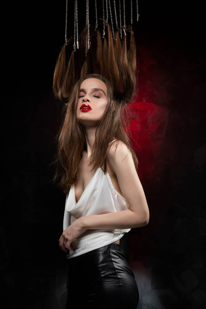 A beautiful girl with evening make-up and red lips wearing a blouse with a deep neckline and her hair suspended on chains, posing on a red and black background. Conceptual, avant-garde design. - Fotoğraf, Görsel