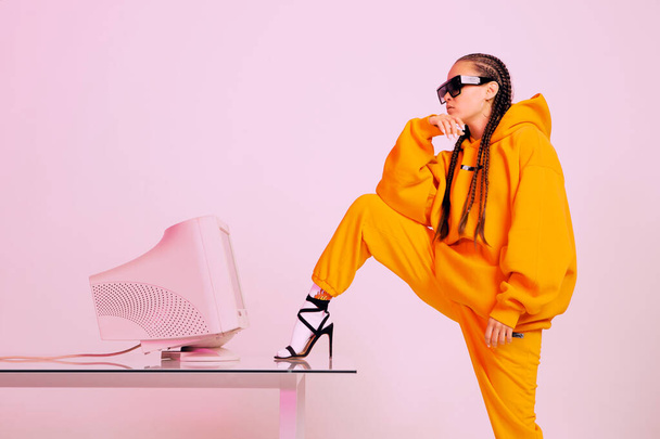 Stylish young female office worker in sunglasses with old computer monitor and foot on desk. Colored light - Photo, image