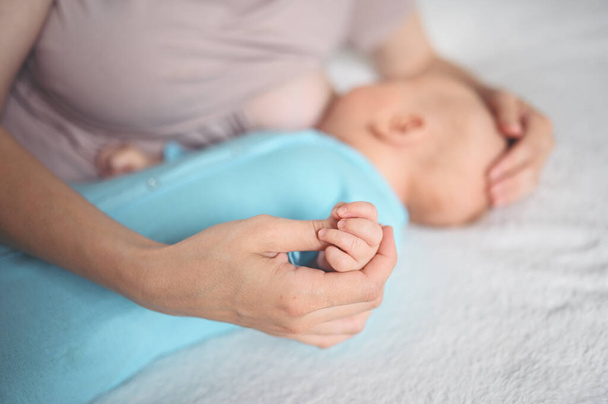 Молодая мать лежит с новорожденным милым младенцем в синем комбинезоне на кровати, обнимает и кормит грудным молоком. Здоровый ребенок, концепция больницы и счастливое материнство. Детская комната. - Фото, изображение