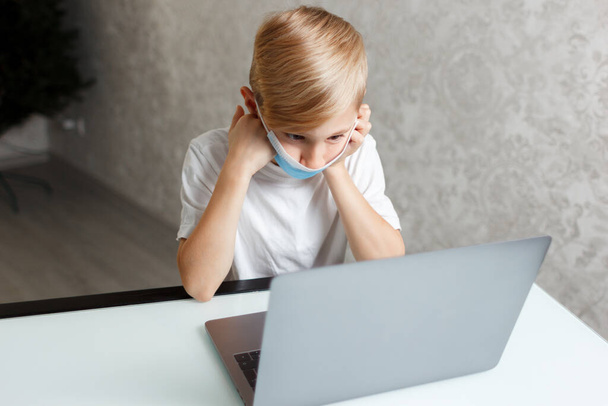 een kind met een medisch masker met een laptop voert huiswerk uit voor online training - Foto, afbeelding