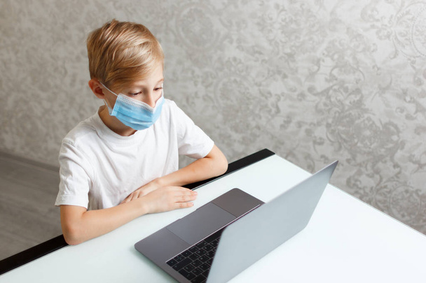 ένα παιδί με ιατρική μάσκα με φορητό υπολογιστή εκτελεί εργασίες για online εκπαίδευση - Φωτογραφία, εικόνα