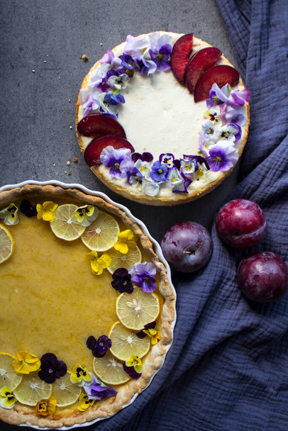 Homemade sweet cakes on a table. Close up photo of lemon tart, cheesecake decorated with flowers and fresh citrus fruits on grey background. Colorful sweet food.  - 写真・画像