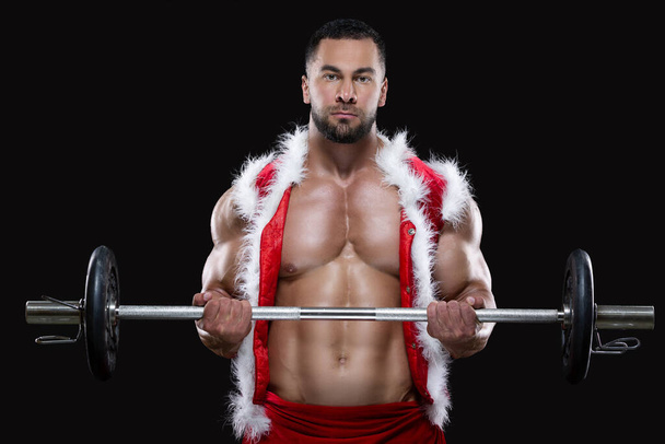 Strong muscular athlete wearing Santa Claus costume over naked body is lifting a barbell isolated on black background - Foto, Imagem