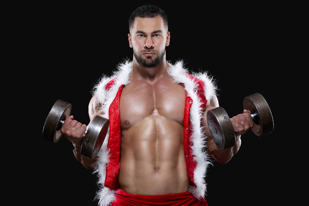 Strong muscular athlete wearing Santa Claus costume over naked body is lifting dumbbells isolated on black background - Foto, Imagem