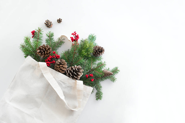 Christmas composition. Eco bag with Christmas tree branches, holly berries and pine cones on white background. Christmas coming and gift delivery concept. Top view, copy space. - 写真・画像