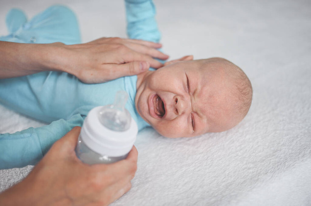 かわいい感情的な泣いて新生児の少年ミルクボトルでベッドの上に横たわっている。赤ちゃんの顔の表情。ベビー用品包装テンプレート。健康な子供,病院と幸せな母親の概念.幼児の赤ちゃん。保育園. - 写真・画像