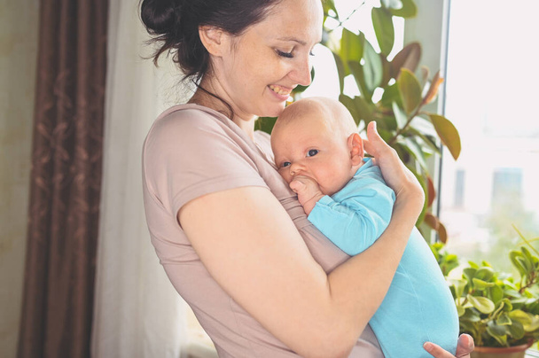 Hermosa madre sosteniendo en brazos a su pequeño y lindo hijo recién nacido. Expresiones faciales de bebé. Familia en casa junto a la ventana. Niño sano, concepto de hospital y maternidad feliz. Vivero. - Foto, imagen