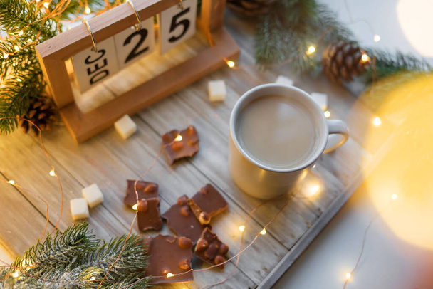 Flatlay coffee with milk and chocolate with hazelnuts on a wooden tray against a background of garlands and fir branches. Christmas dessert on December 25 with lights in bokeh. - Photo, Image