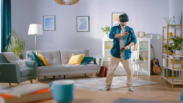 Jeune homme énergique portant un casque de réalité virtuelle et tenant des contrôleurs joue dans un jeu vidéo à la maison. Homme jouant VR Shooter au milieu du salon. - Photo, image