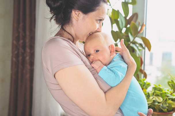 Piękna matka trzymająca w ramionach swojego małego, słodkiego noworodka. Wyraz twarzy dziecka. Rodzina w domu przy oknie. Zdrowe dziecko, koncepcja szpitala i szczęśliwe macierzyństwo. Przedszkole. - Zdjęcie, obraz
