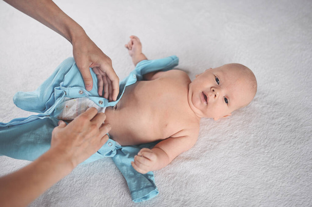 Η μαμά ντύνεται χαριτωμένα συναισθηματικά αστεία νεογέννητο αγοράκι με μπλε φόρμα. Πρότυπο συσκευασίας ειδών μωρών. Υγιές παιδί, έννοια του νοσοκομείου και ευτυχισμένη μητρότητα. Βρέφος. Φυτώριο. - Φωτογραφία, εικόνα